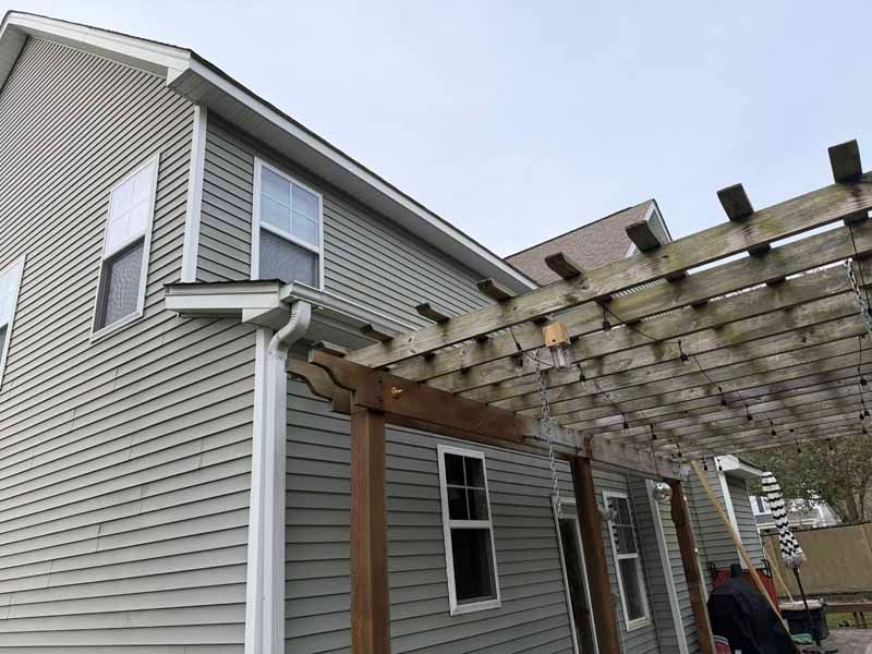 Gray house with a wooden pergola attached. A person stands nearby.
