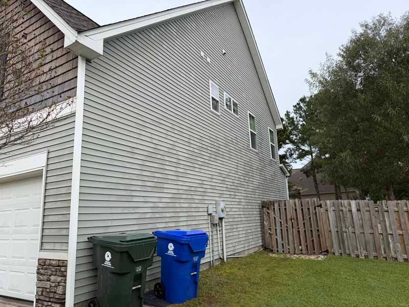 Side of a light gray house with a garage, siding, and a wooden fence. Two trash bins are in front.