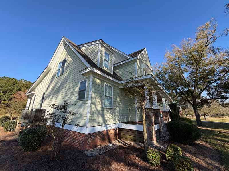 Two-story light green house with white trim, red brick foundation, and a porch, set in a sunny, grassy field.