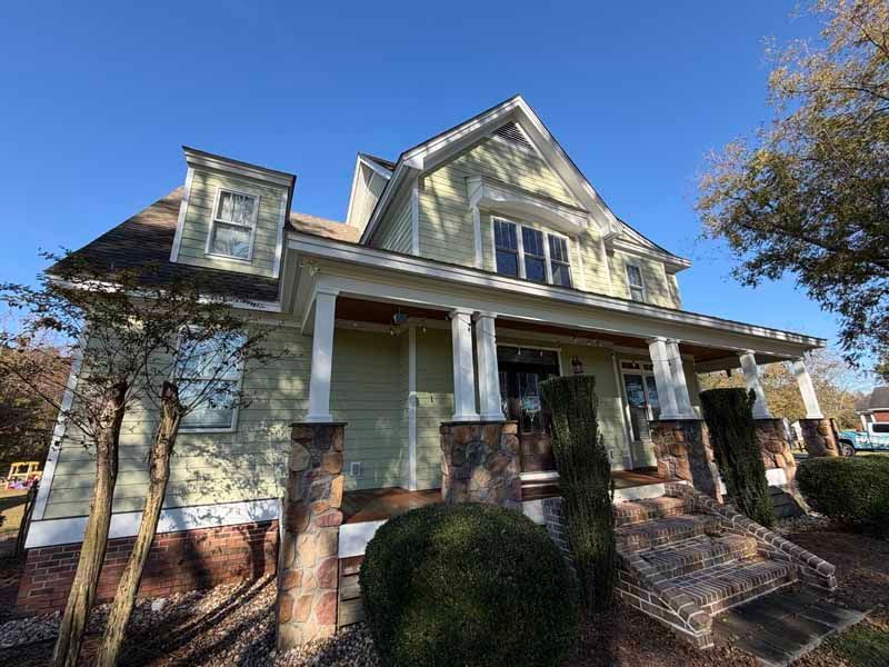 Two-story house with green siding, brick accents, and a front porch under a clear blue sky.
