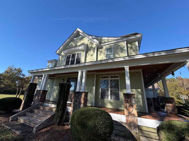 Two-story house with green siding, white trim, porch, and stone accents against a bright blue sky.