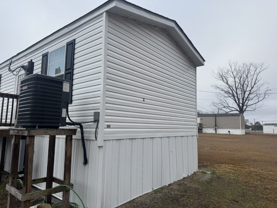 White mobile home with black AC unit on a wooden structure, gray sky, and a bare tree.