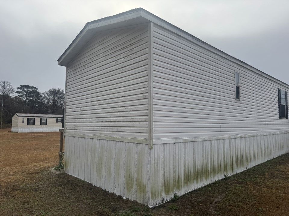 White mobile home with mold on lower siding, another in background, cloudy sky.