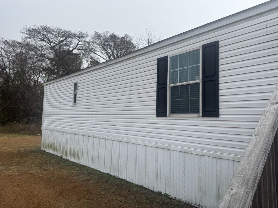 White mobile home exterior with dark shutters, mossy skirting, and bare trees.