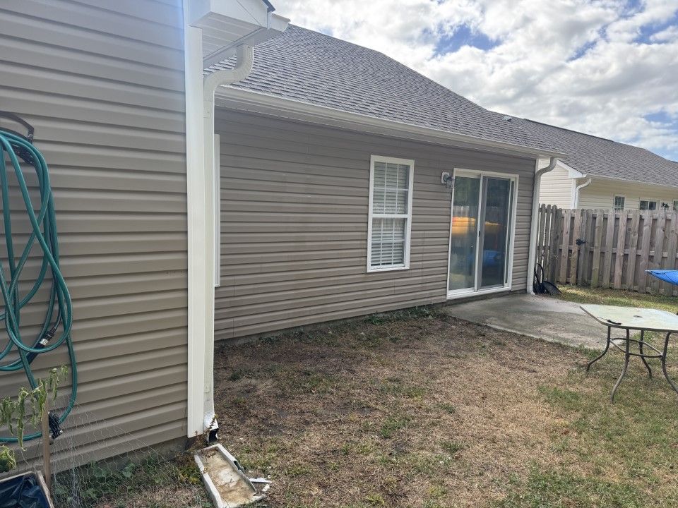 Exterior of a house with gray siding, window, and sliding glass door. A concrete patio and dry grass are in the yard.