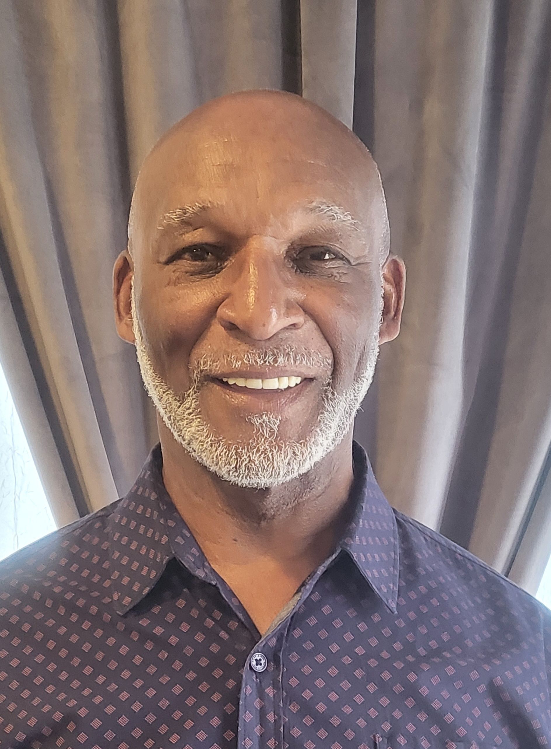 Smiling man in a dark patterned shirt standing before gray curtains.