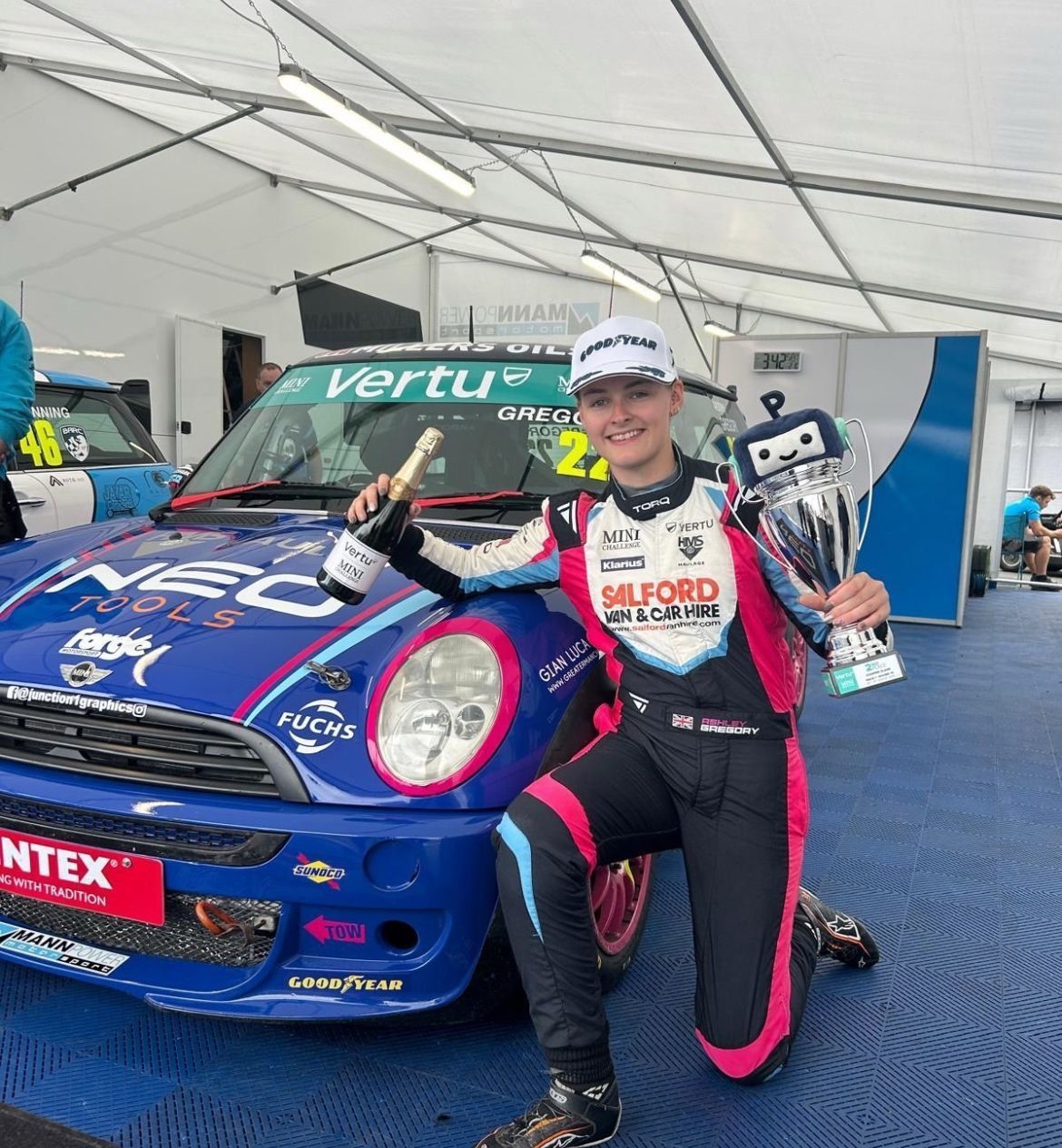 A young woman in racing gear kneels by a blue Mini Cooper, holding a trophy and champagne, celebrating a victory.