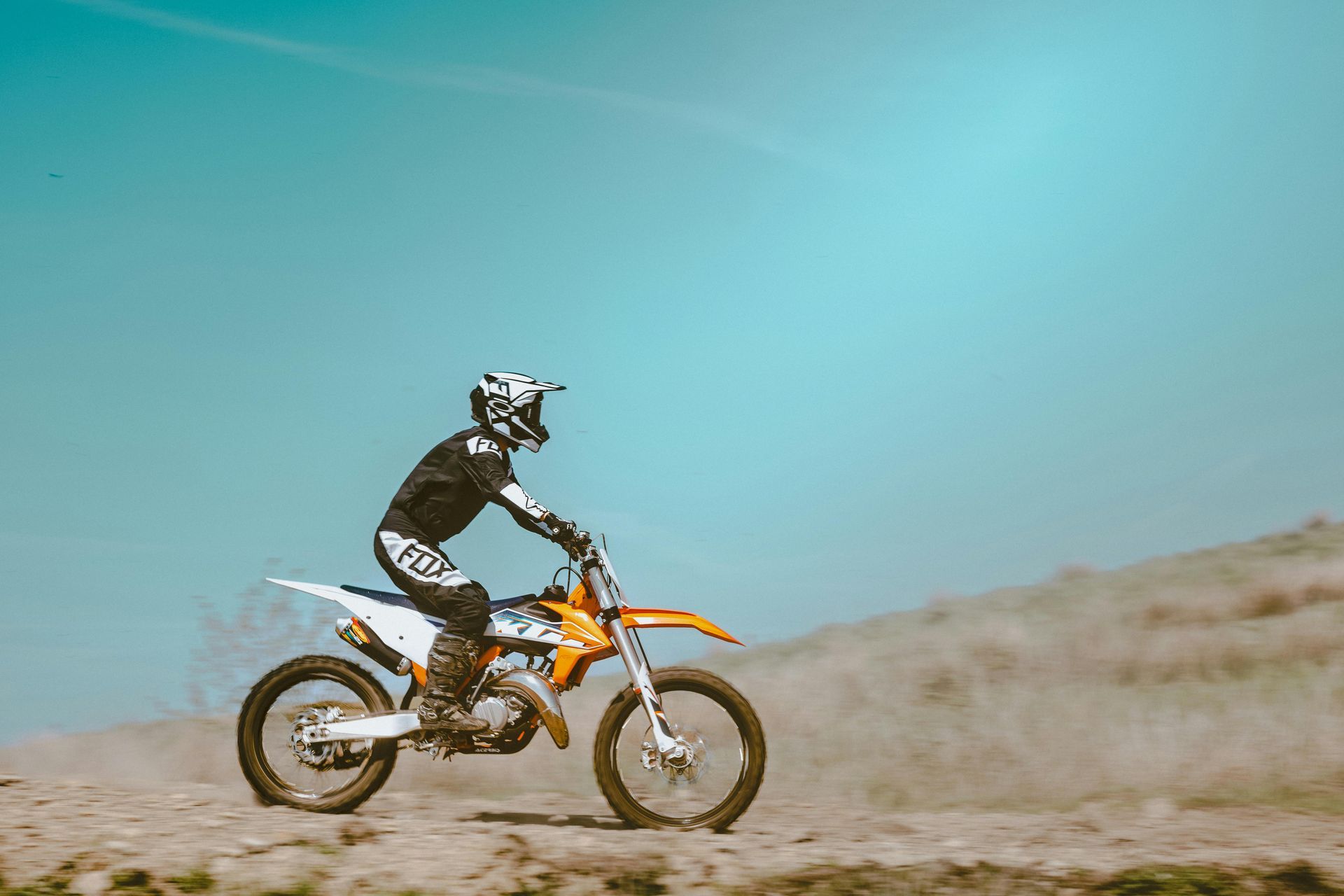 Person on a dirt bike kicking up dust, riding across a dirt track under a blue sky.