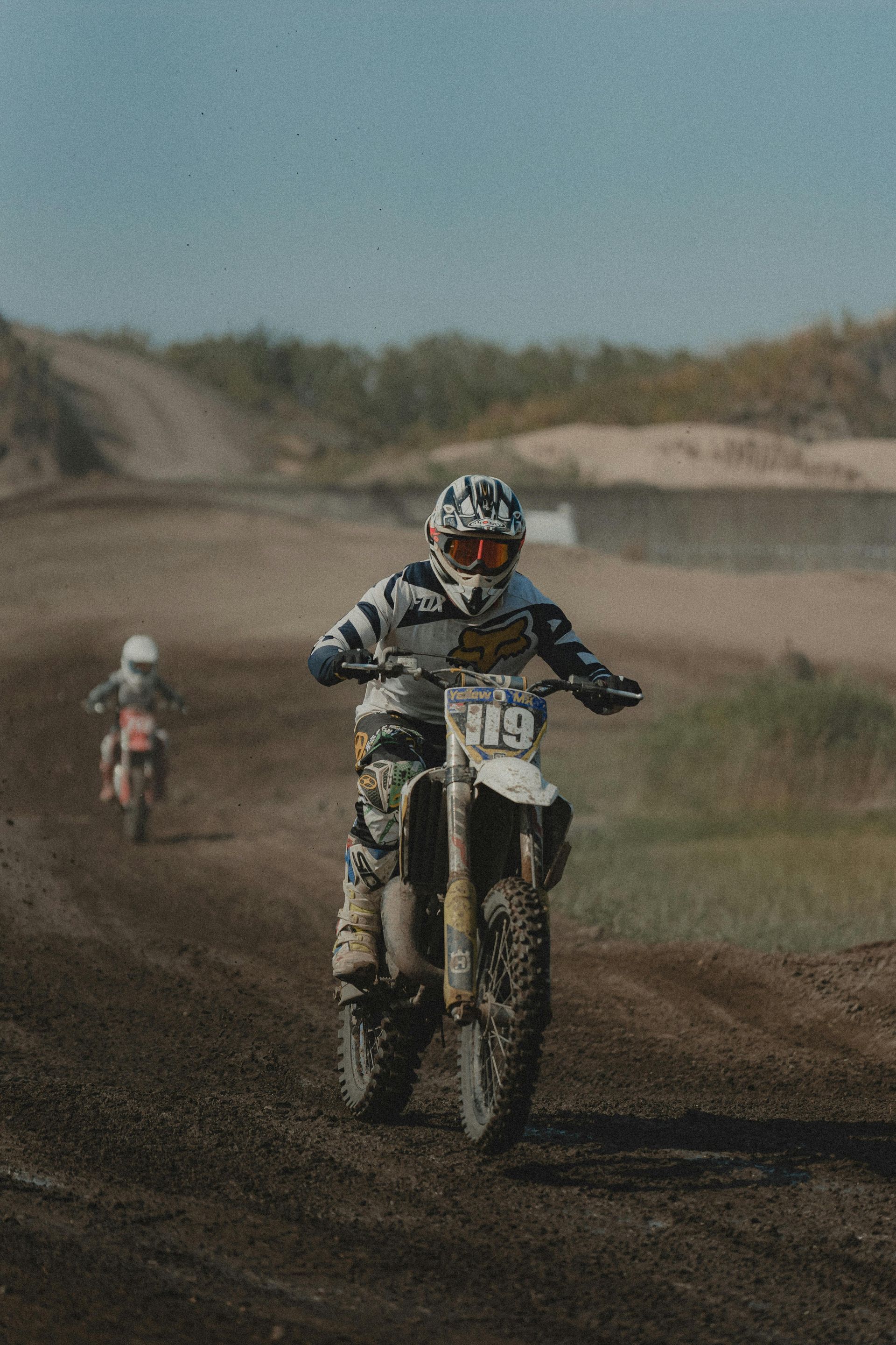 Dirt bike racer on a muddy track; another racer follows in the distance, racing outdoors.