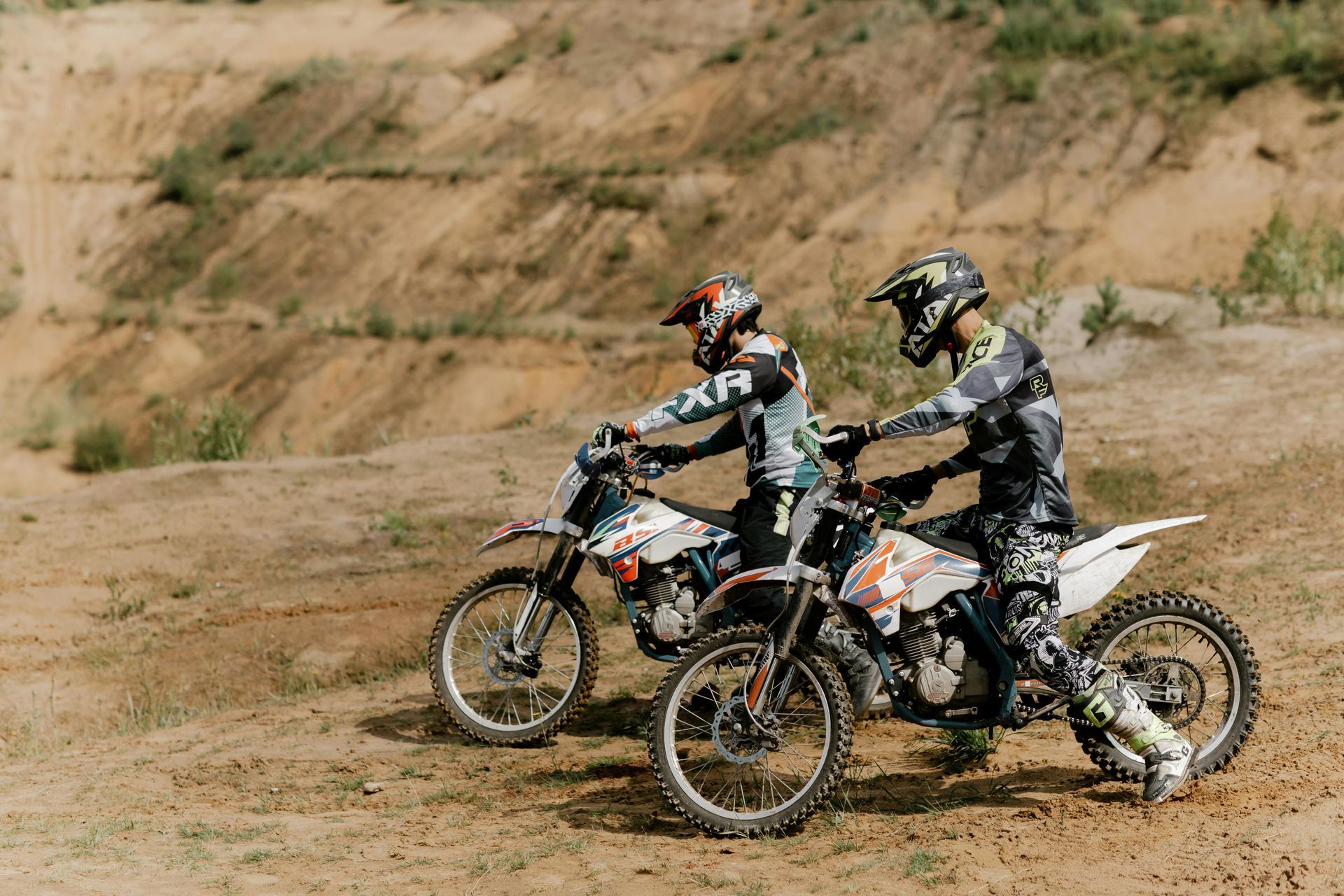 Two motocross riders on dirt bikes on a dirt track, wearing helmets and gear.