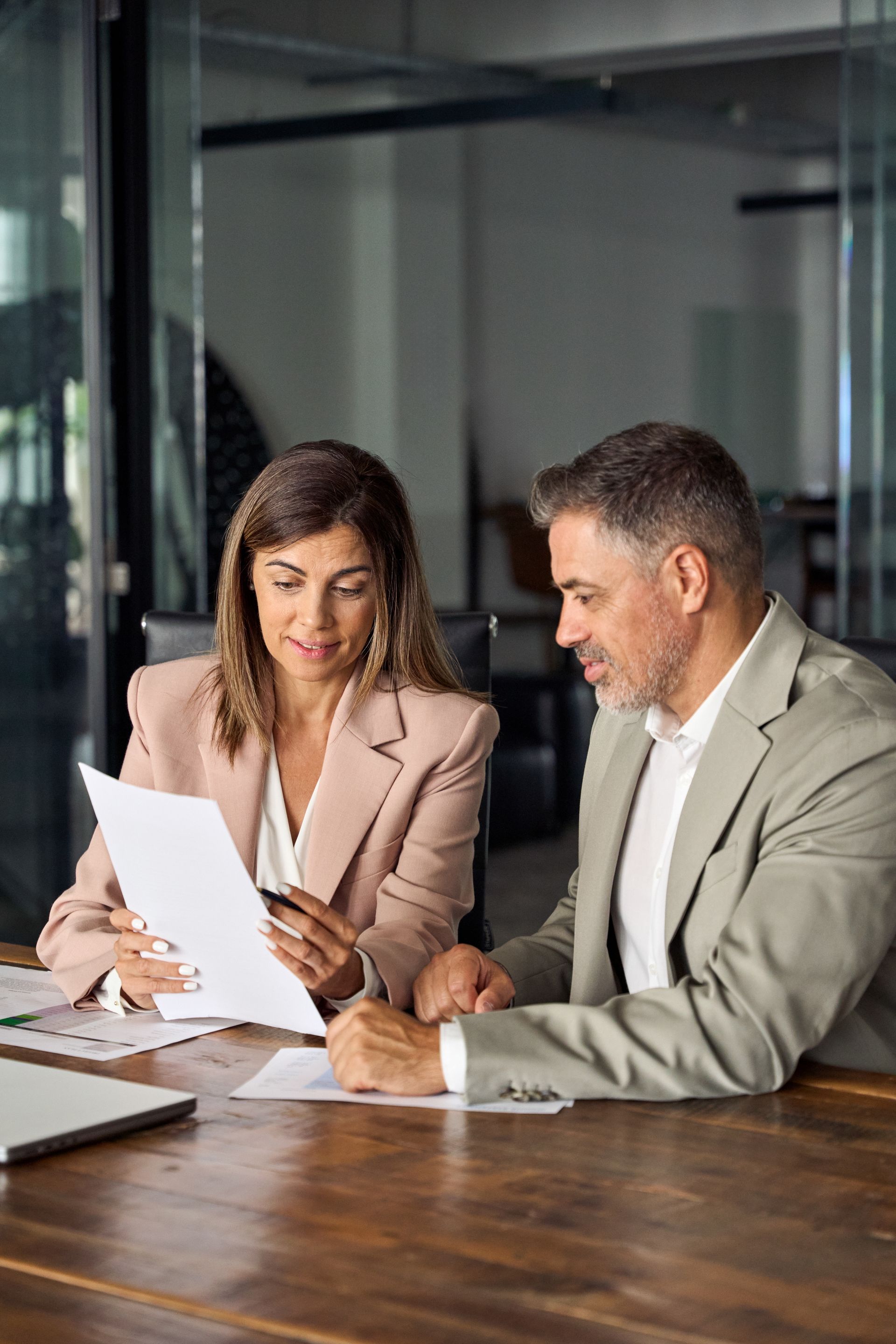 Two professionals reviewing documents together at a wooden table in a glass-walled office