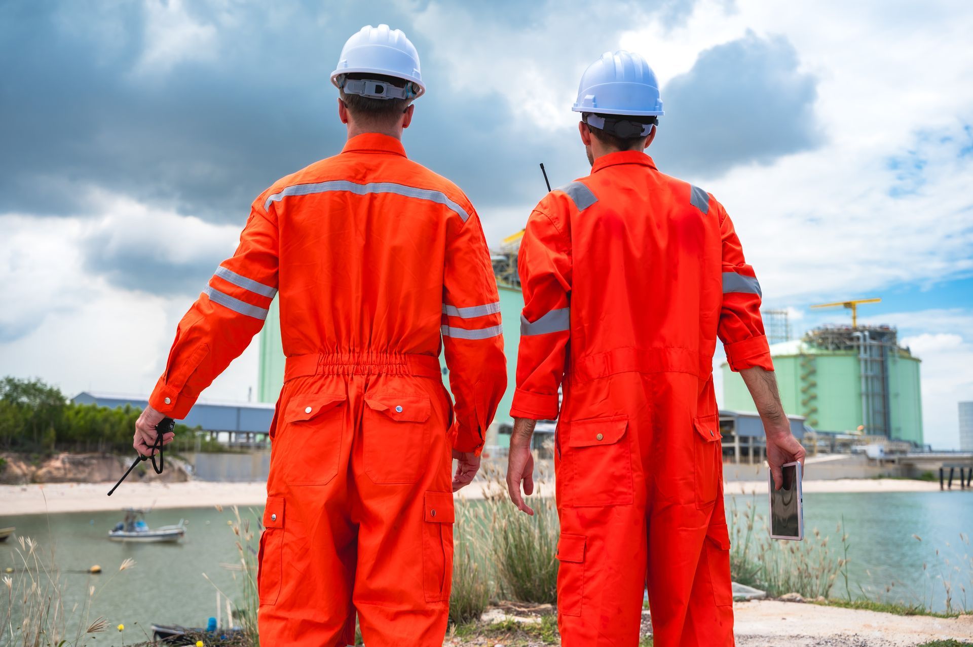 Two workers in orange coveralls and hard hats stand by a waterfront construction site.