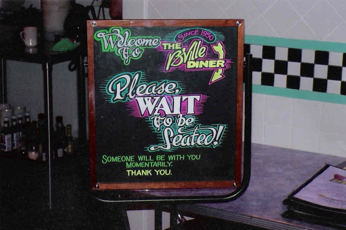 A chalkboard in a diner that says please wait to be seated