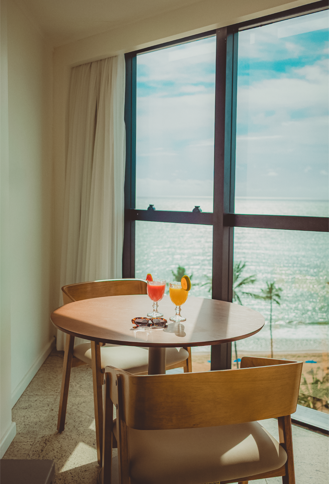 Uma pequena mesa com dois coquetéis e vista para o oceano através de uma grande janela.