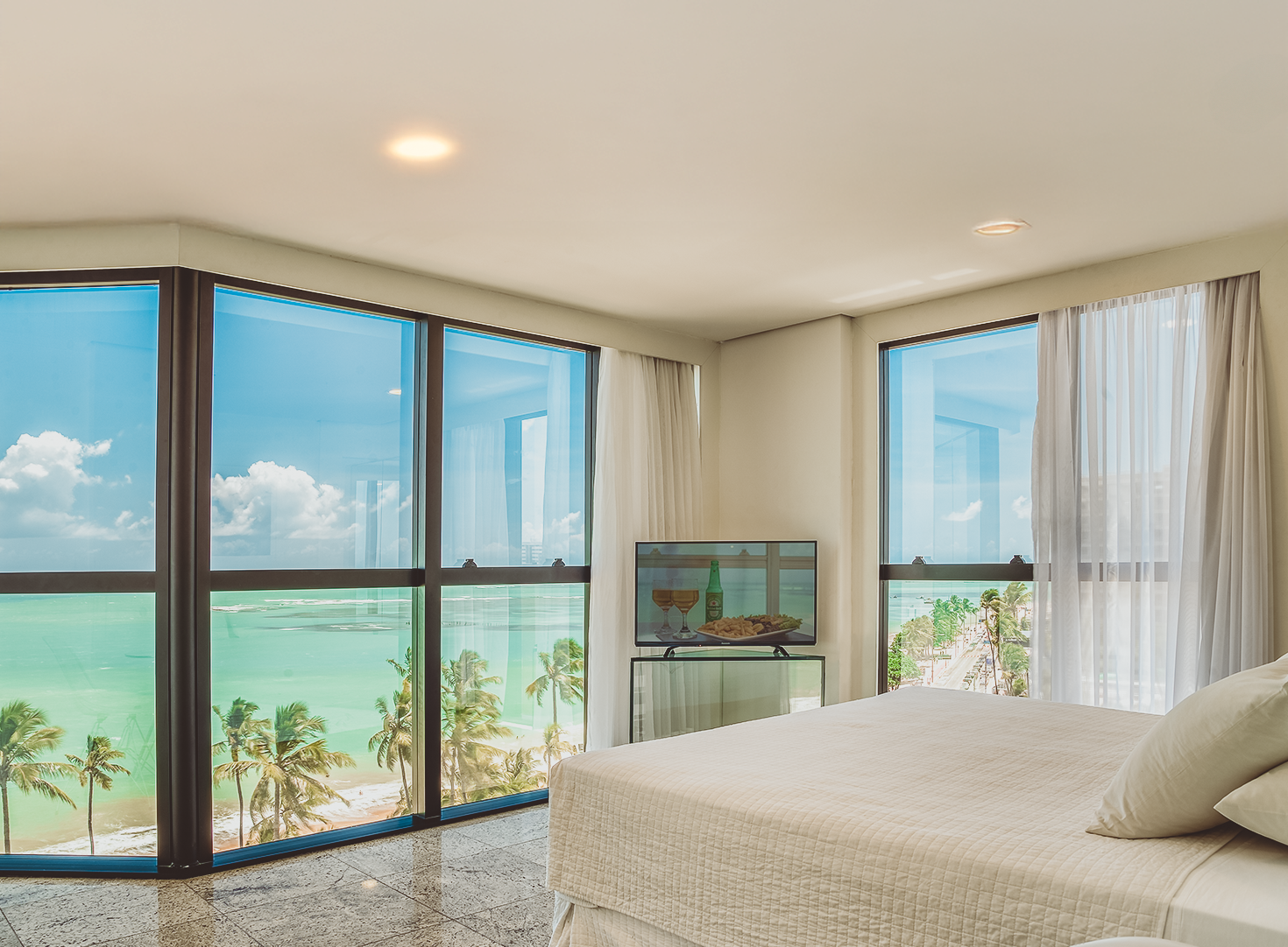 Quarto com vista para o mar, janelas grandes, cama com lençóis brancos, TV e cortinas brancas.