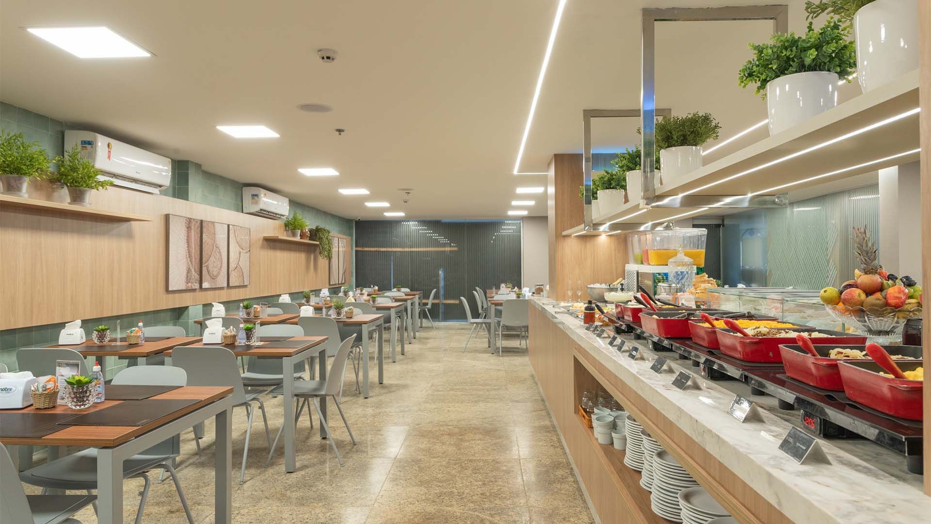Buffet de café da manhã do hotel com mesas e exposição de alimentos; cores neutras, ambiente interno.
