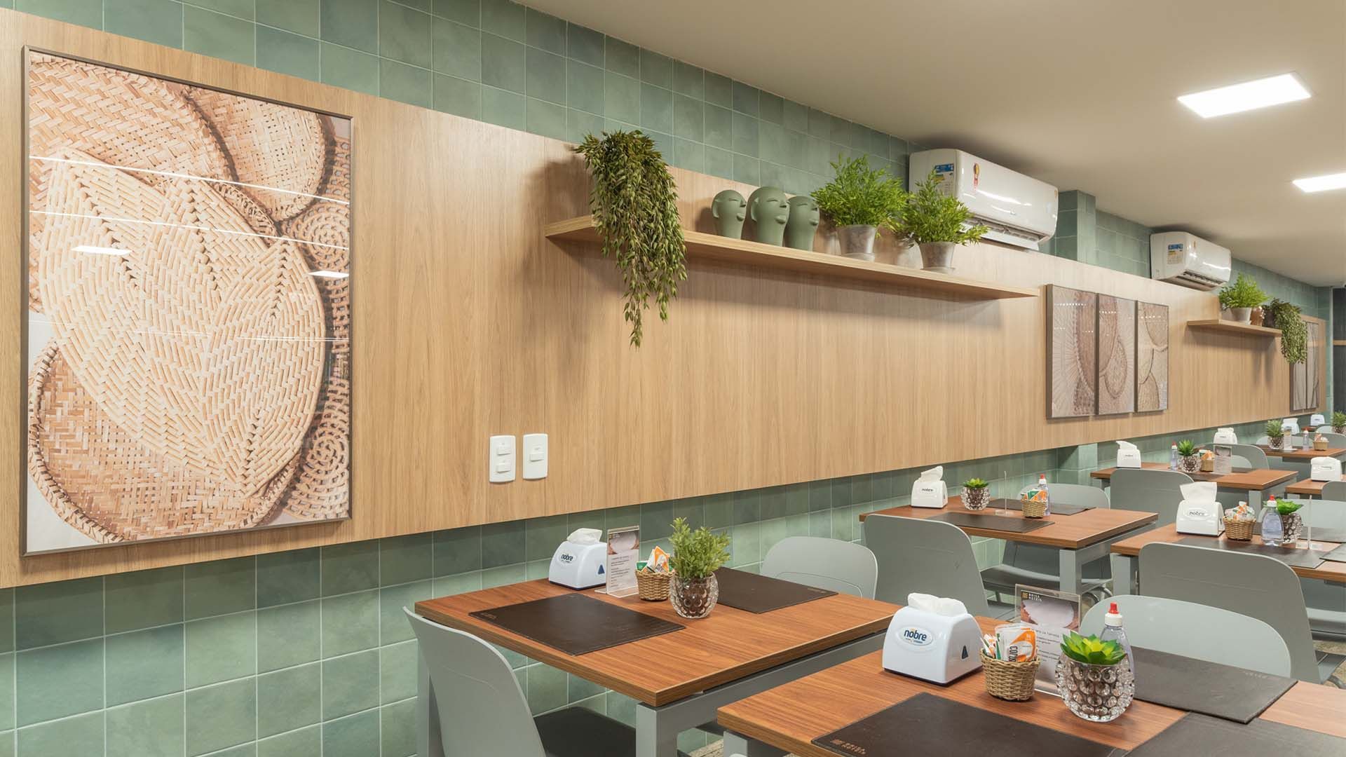 Interior do restaurante com painéis de madeira, plantas, mesas quadradas, cadeiras cinzas e uma obra de arte decorativa com folhas.