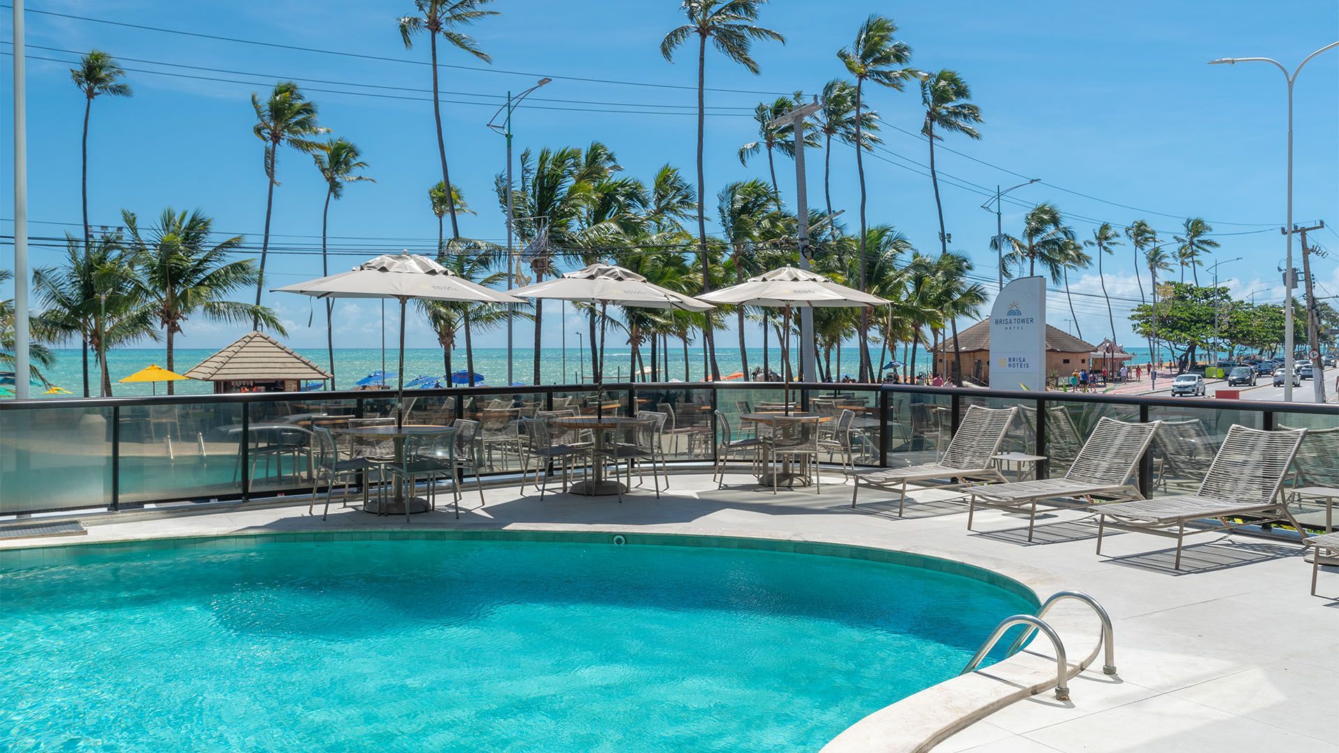 Piscina com espreguiçadeiras, mesas e guarda-sóis perto da praia em um ambiente ensolarado.