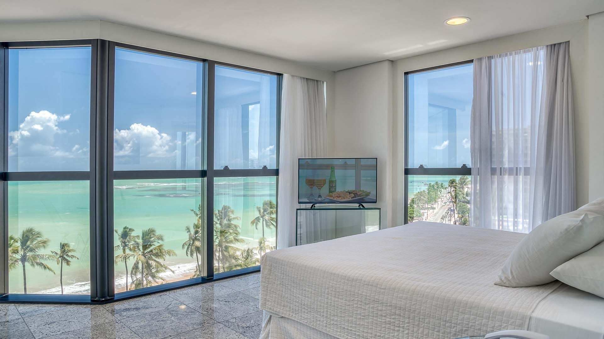 Quarto com janelas amplas com vista para uma praia com palmeiras e águas azul-turquesa. Cama branca e TV.