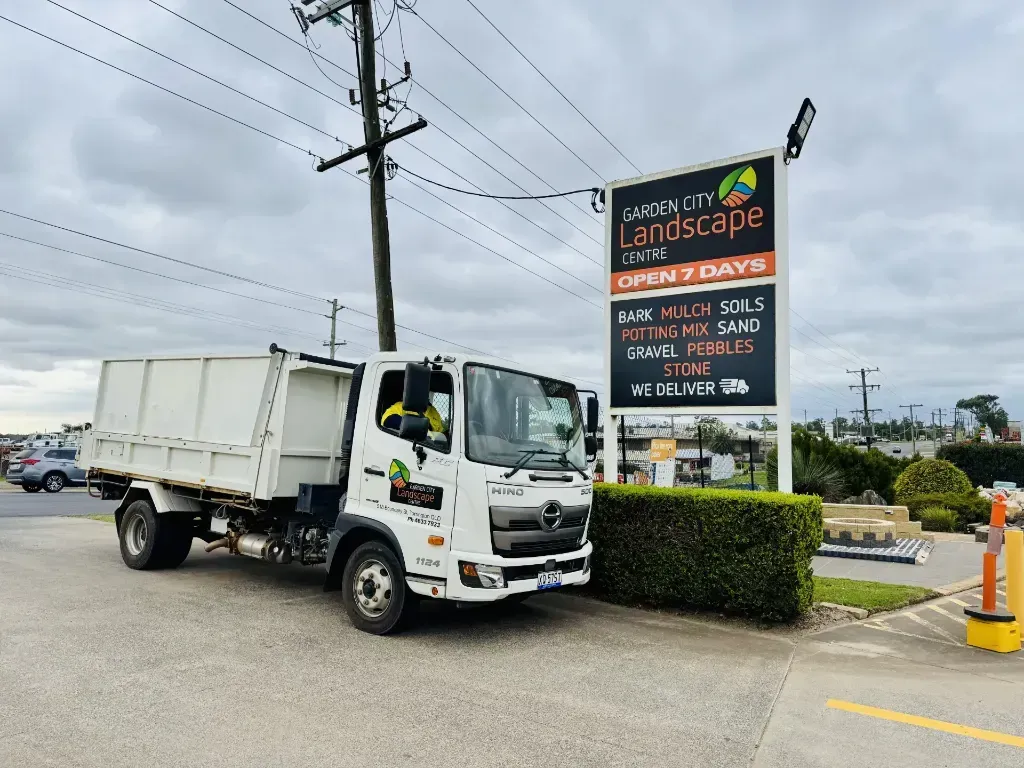 A dump truck is parked in front of a sign that says garden city landscape centre is open 7 days.