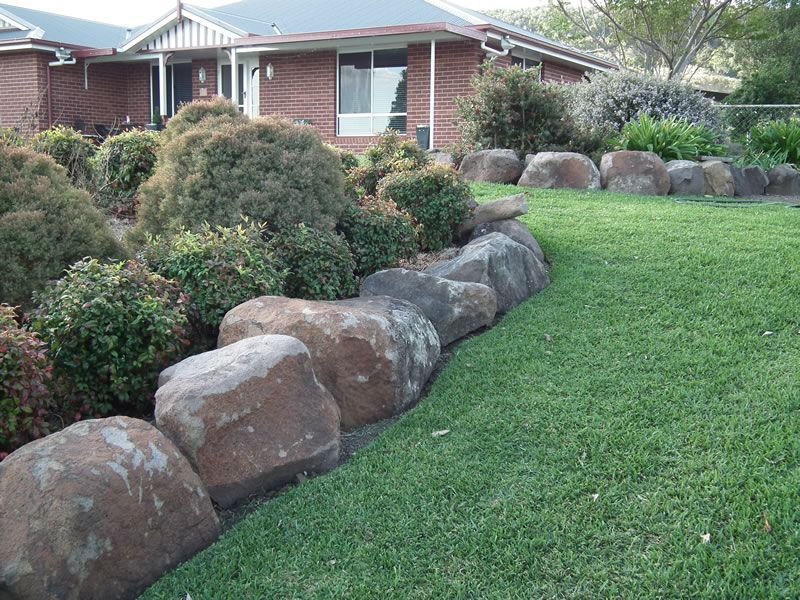 A large rock wall surrounds a lush green lawn in front of a brick house.