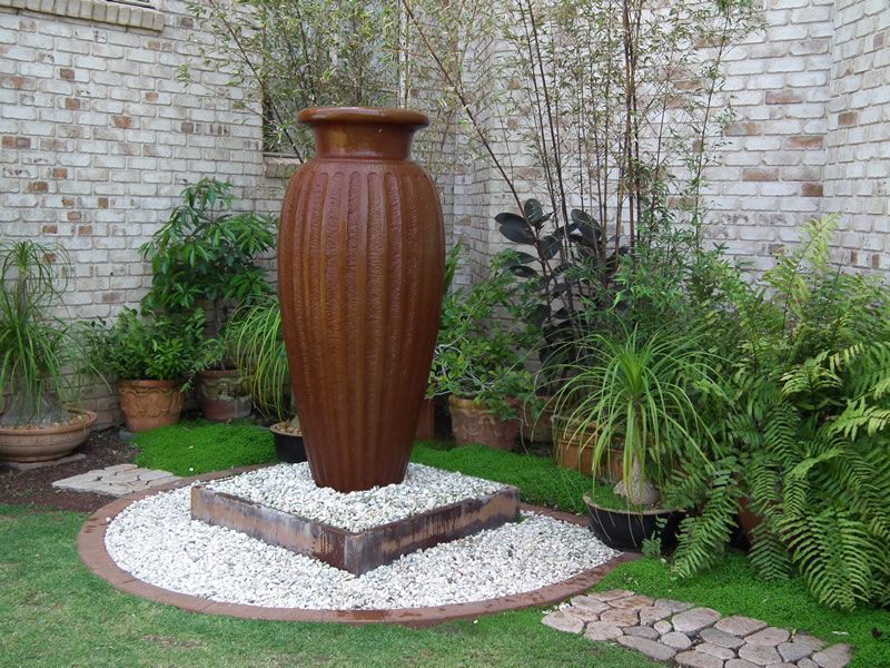 A large vase is sitting in the middle of a garden surrounded by potted plants.