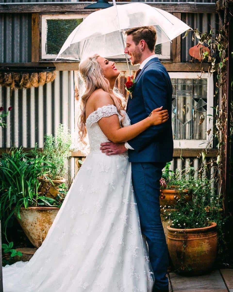 A bride and groom are standing under an umbrella in the rain — Glam Hair and Beauty in Woy Woy, NSW
