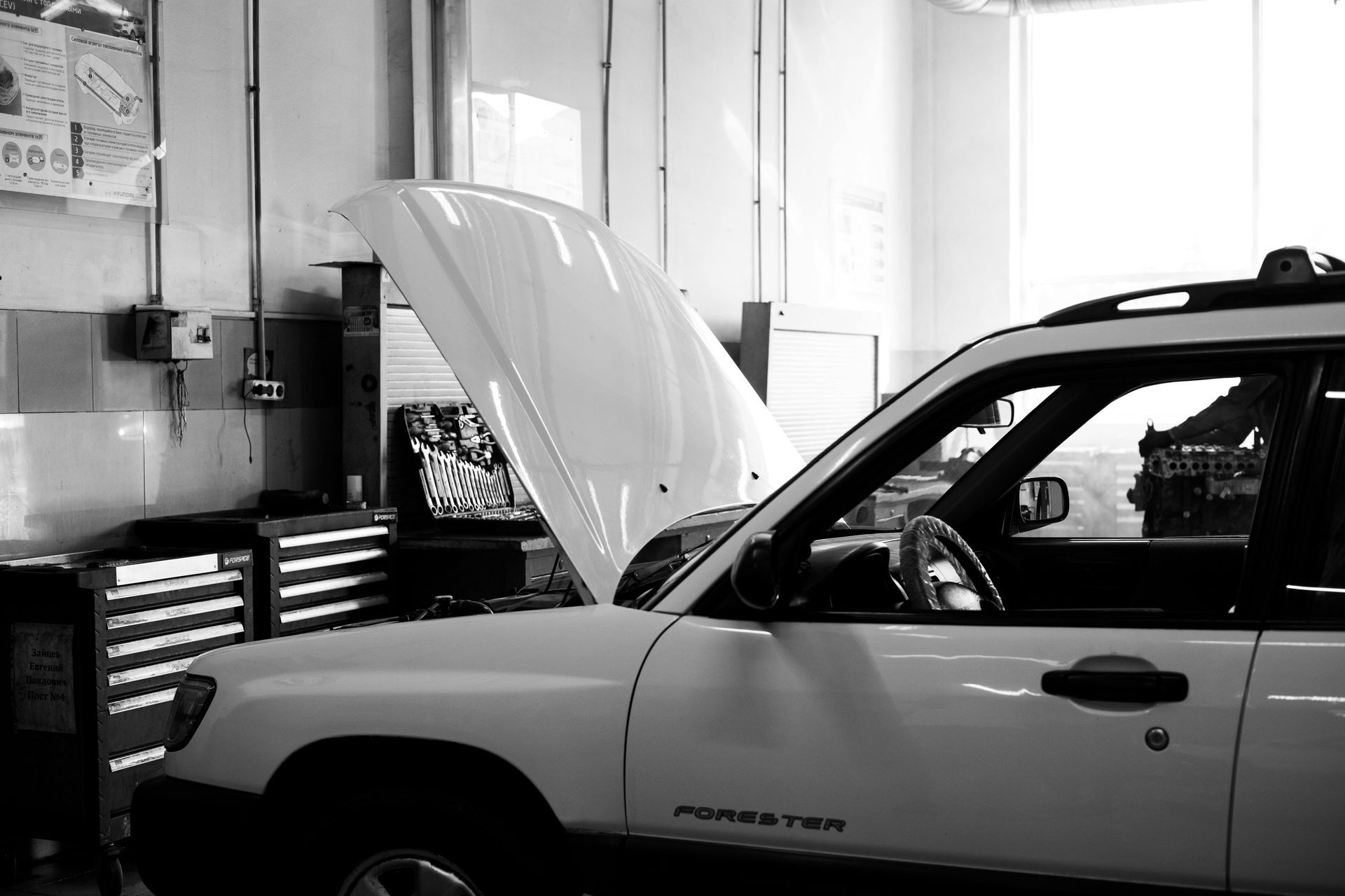 Car with hood open in a mechanic’s garage, next to tool cabinets and workshop equipment.