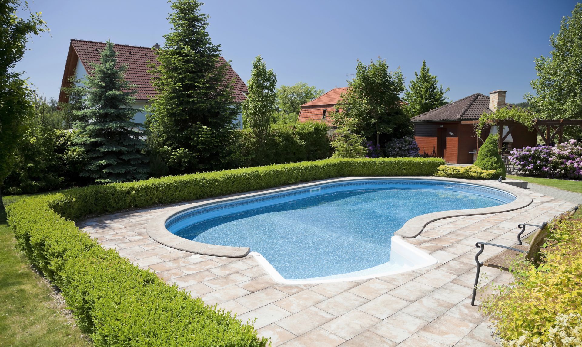 Piscina in giardino con acqua azzurra, patio in pietra, siepi e alberi in una giornata di sole.