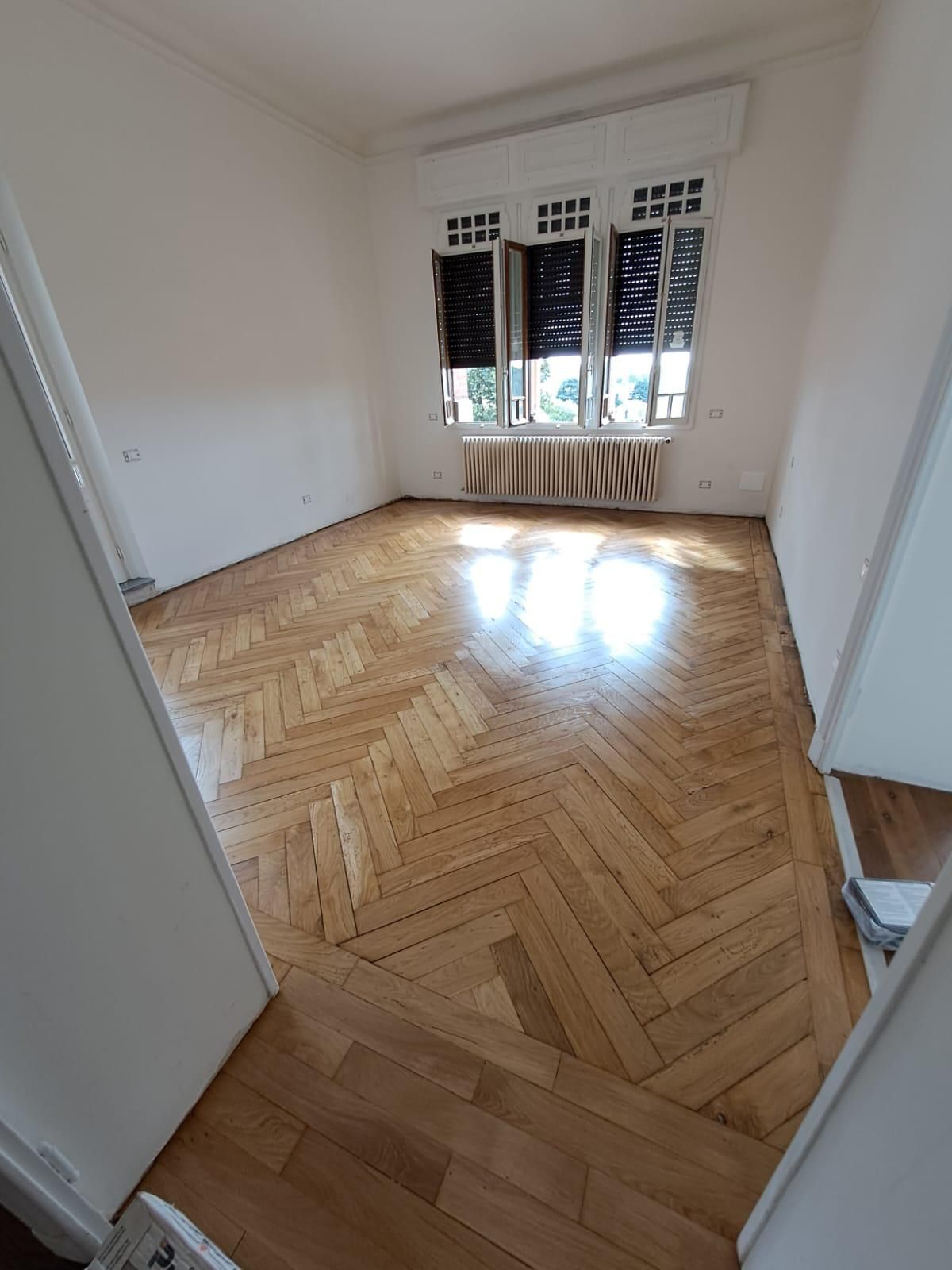 Stanza bianca vuota con pavimento in legno a spina di pesce e luce intensa proveniente dalla finestra.