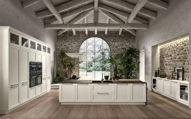 Cucina moderna e luminosa con mobili bianchi, isola centrale, finestra ad arco e travi in ​​legno a vista.