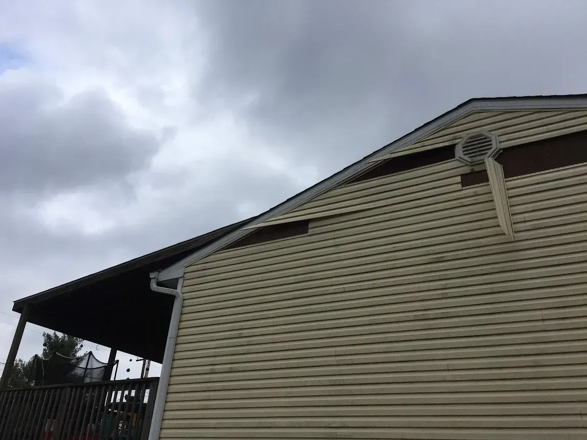 Beige vinyl-sided house with brown trim and dark porch roof against a cloudy sky.