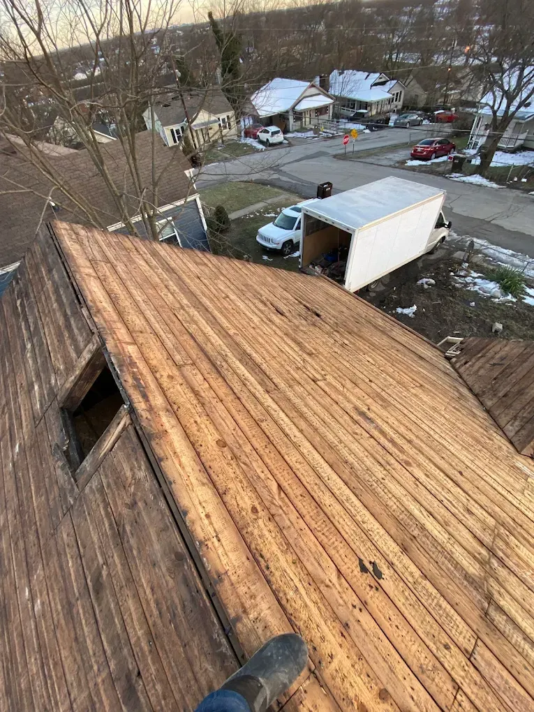 Overhead view of a weathered wooden roof with a small window, a white truck and snowy neighborhood in the background.