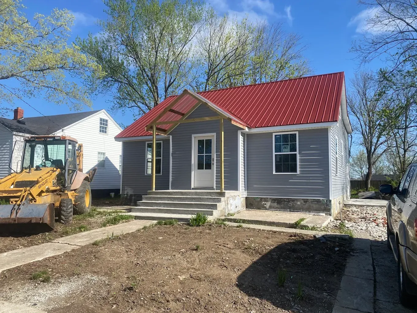 Gray house with a red roof under construction; a yellow excavator is in the yard.