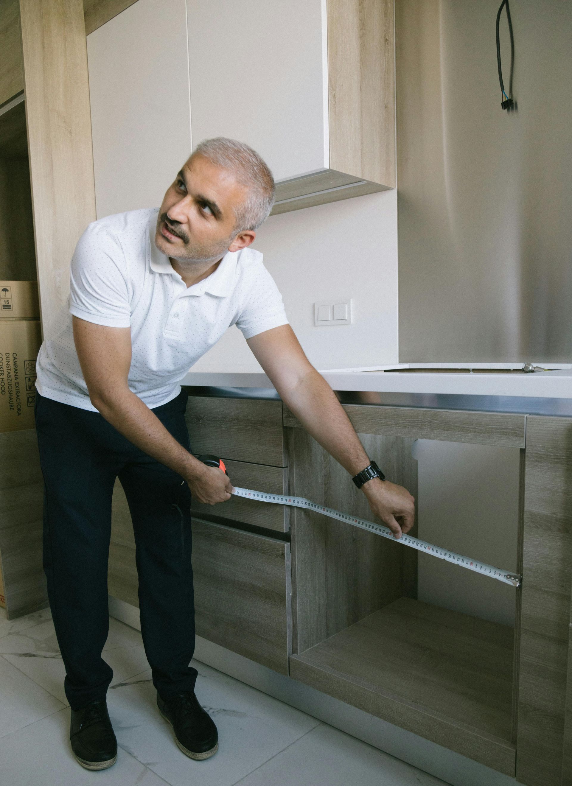 Man measuring inside of a kitchen cabinet; neutral colors, white shirt, dark pants, looks up.