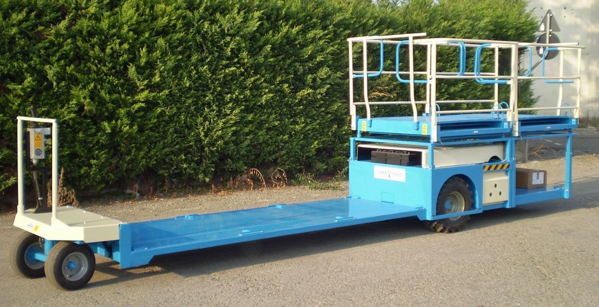 Carrello industriale blu e bianco con piattaforma e ringhiera, parcheggiato sul marciapiede, con cespugli verdi sullo sfondo.