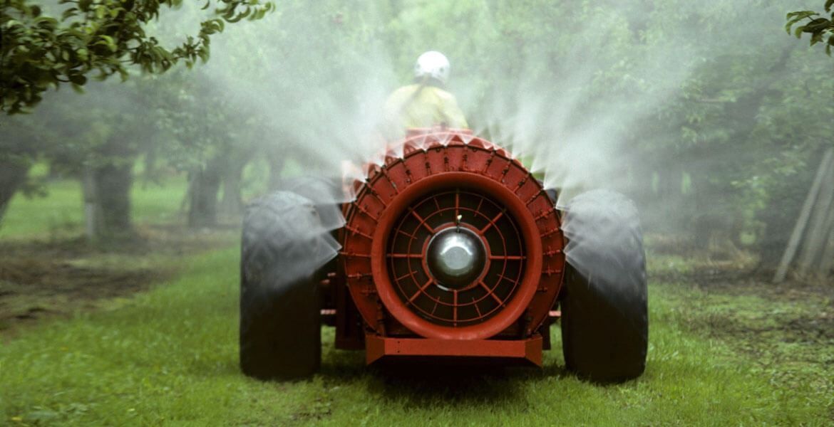 Trattore rosso che irrora le colture in un frutteto; la macchina è circondata da nebbia d'acqua.