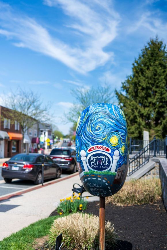 Painted blue face sculpture with vivid swirls on a post beside a sidewalk and parked cars