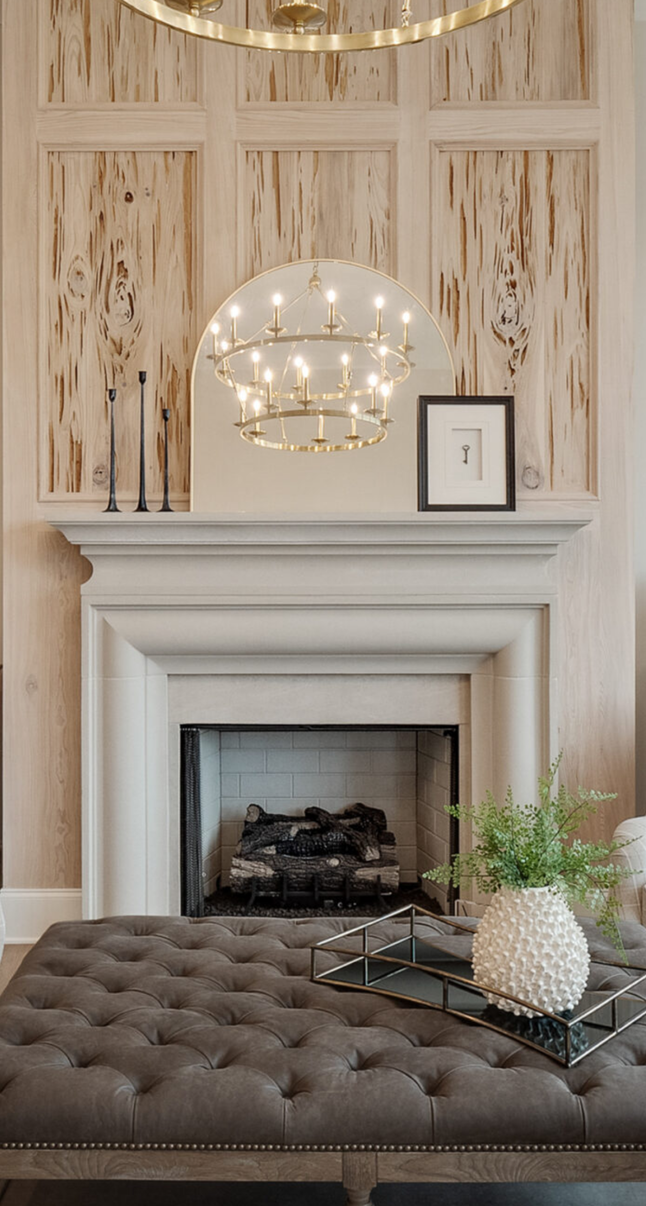Fireplace with white mantel, a tufted ottoman, and a decorative chandelier.