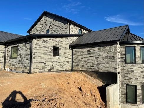 Stone-clad house with dark metal roof, black gutters and windows. Built on a hillside, clear blue sky.