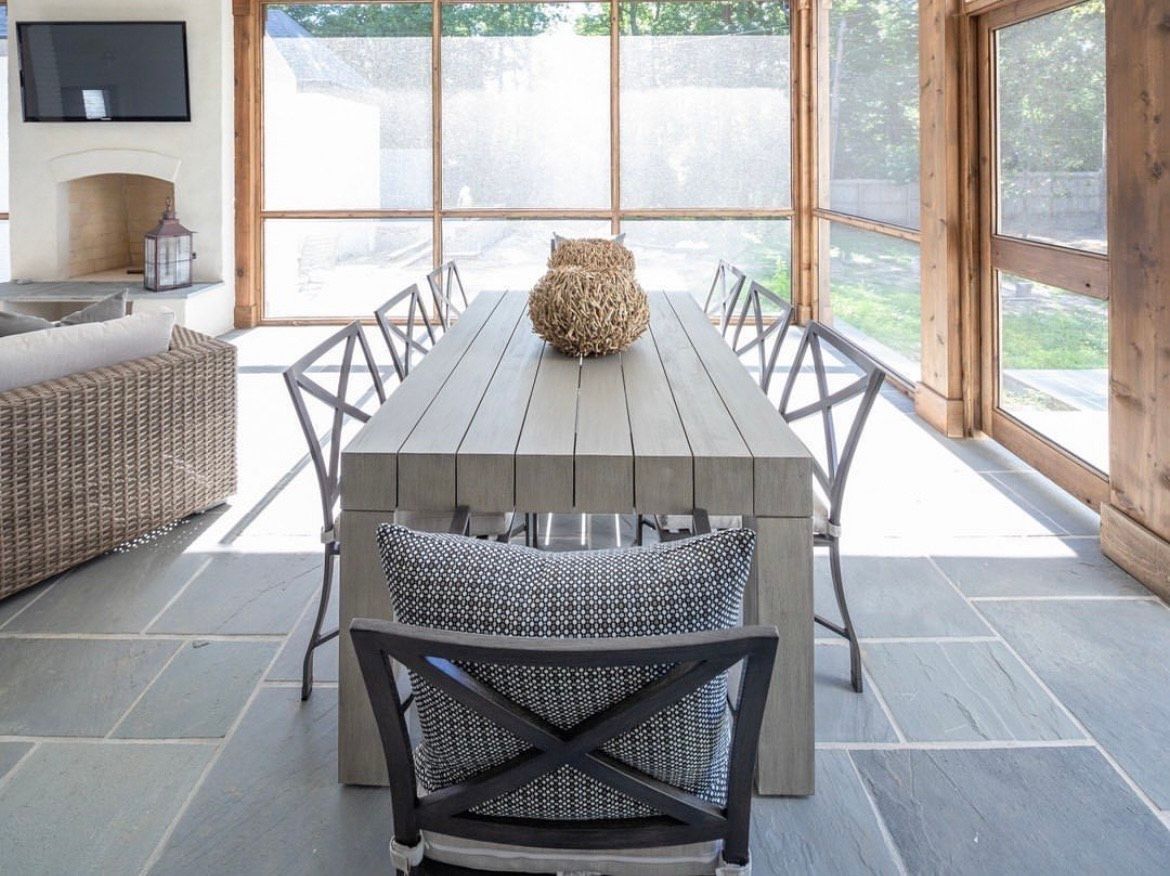 Outdoor dining area with long gray table, chairs, and stone tile floor.