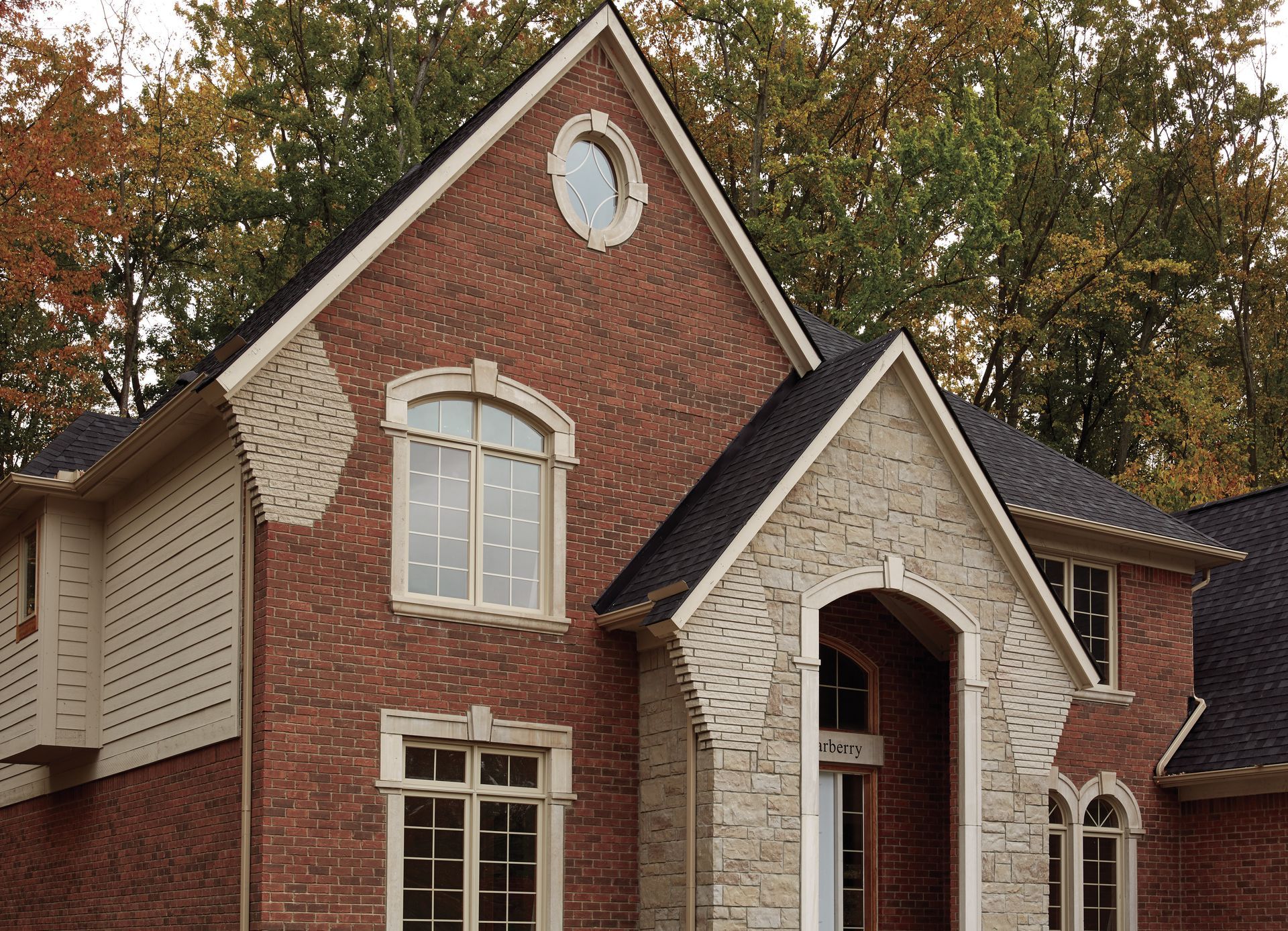 Two-story brick house with a gabled roof, tan siding, and arched windows. Fall trees in the background.