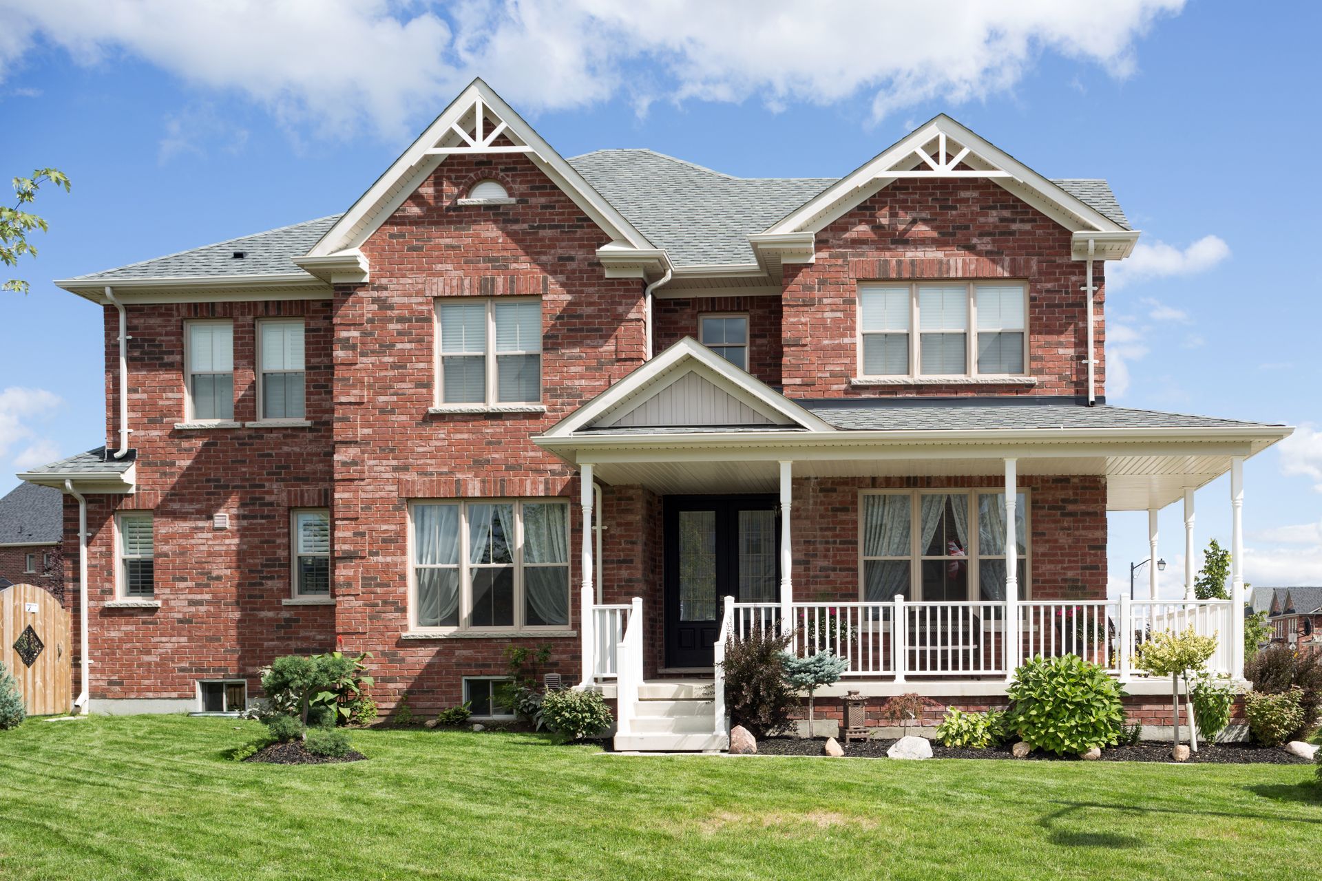 Two-story brick house with a front porch and a green lawn under a blue sky.