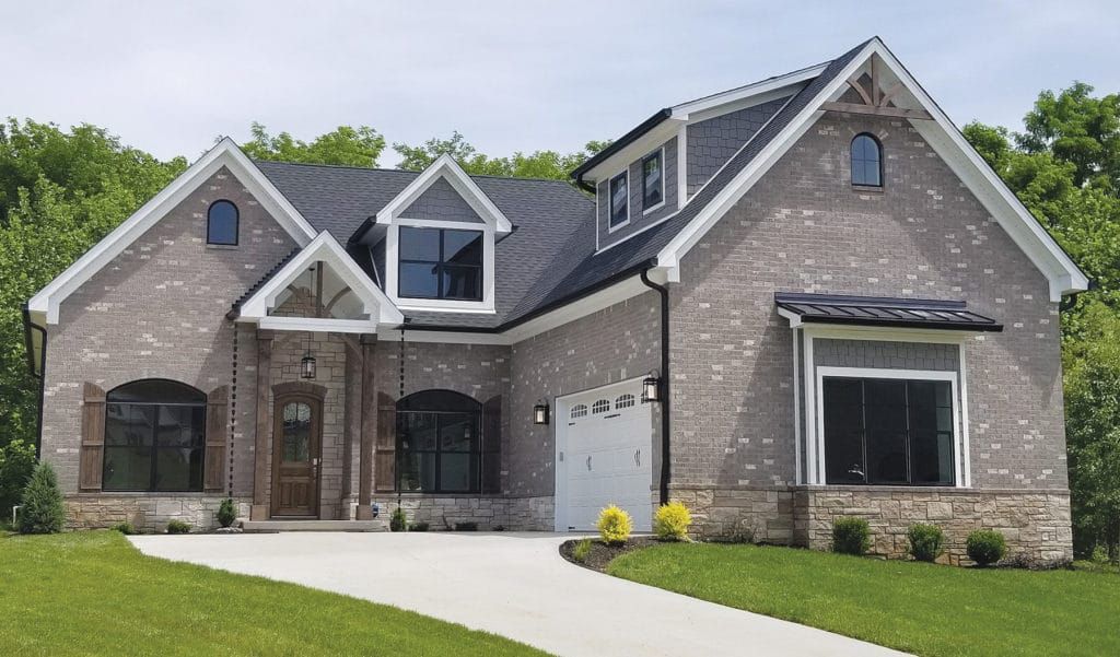 Brick-faced house with a gray roof, dormers, and a white garage door, with a paved driveway and green lawn.
