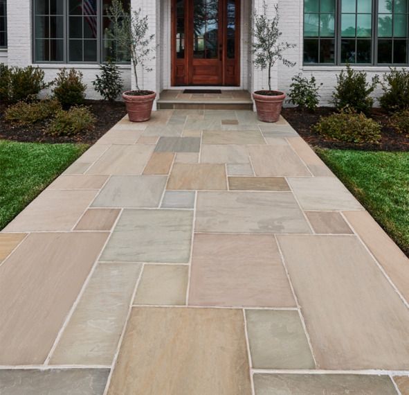 Stone walkway leading to a house entrance with brown door and potted trees. Green grass borders the walkway.