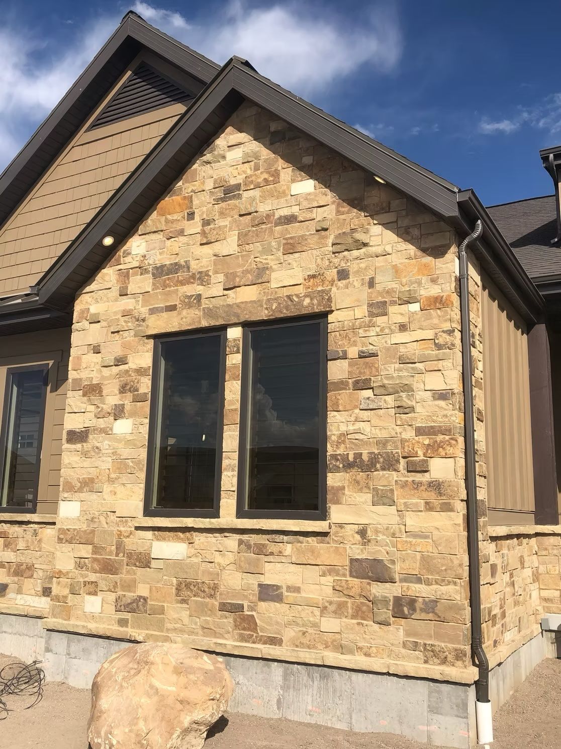 Stone-clad house exterior with dark-framed windows, brown trim, and a blue sky.