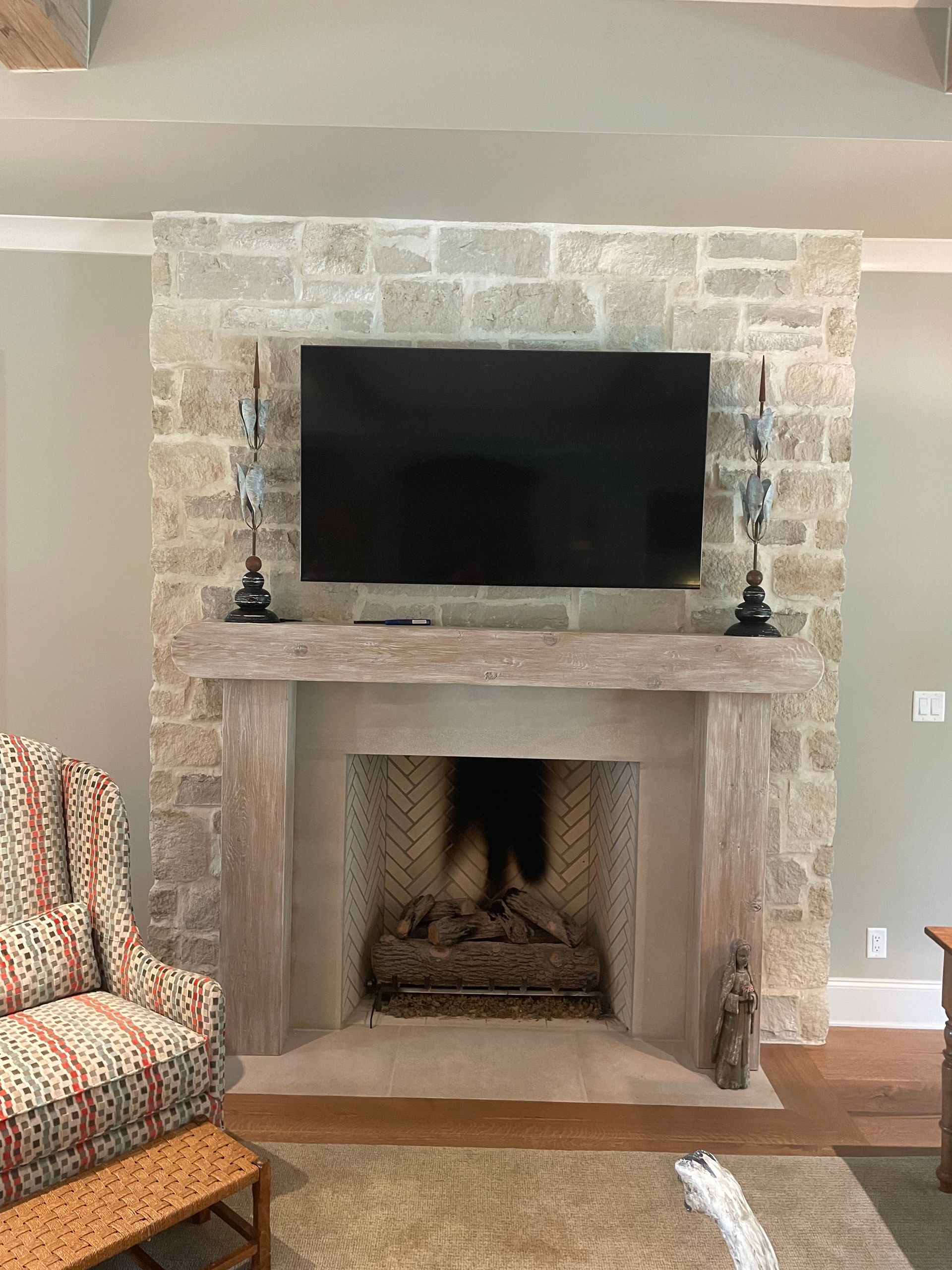 Fireplace with stone surround, TV, mantle, and decorative items. Chair visible in foreground.