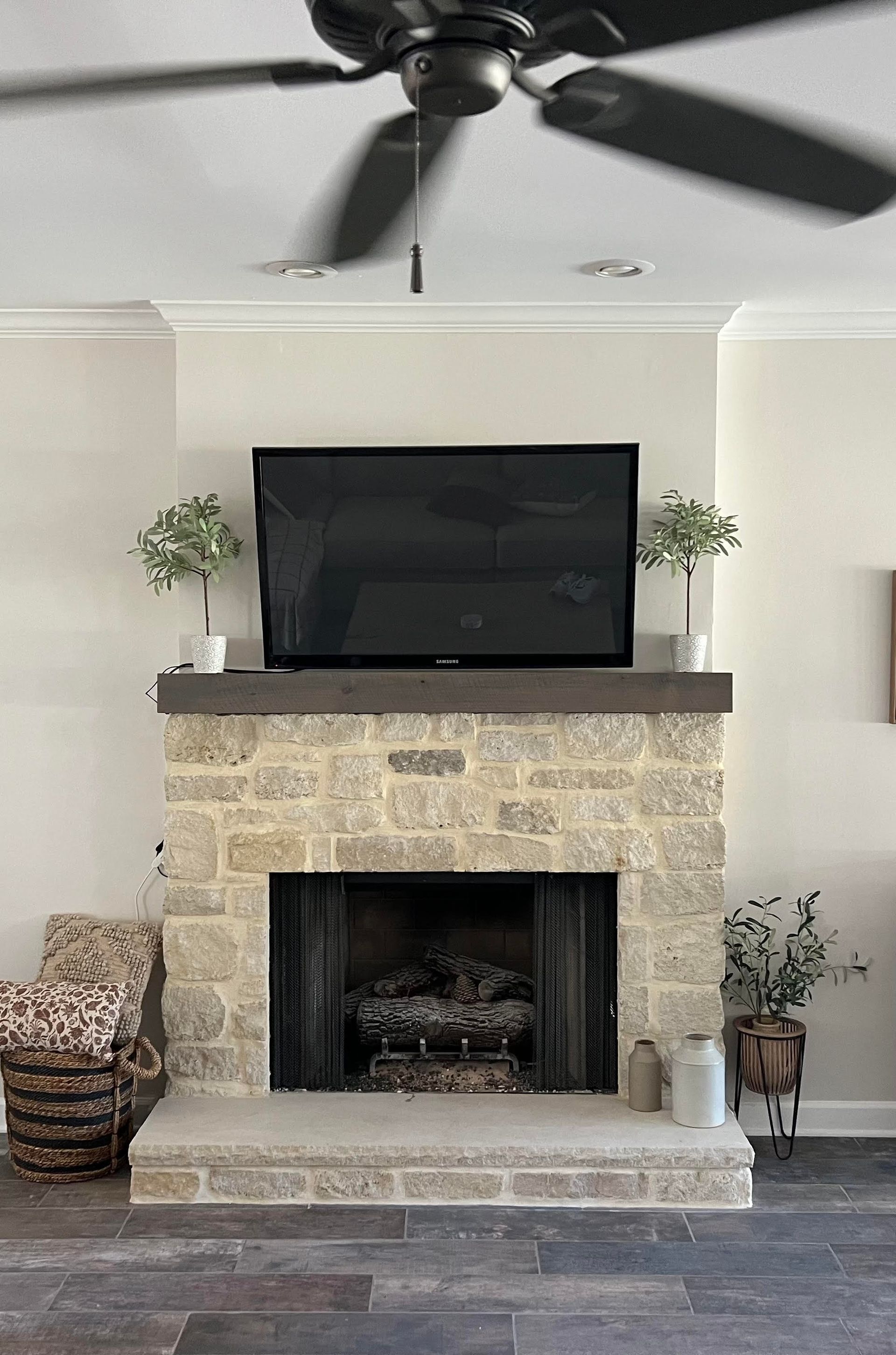 Fireplace with stone facade, dark TV above, plants on mantle. Dark ceiling fan.