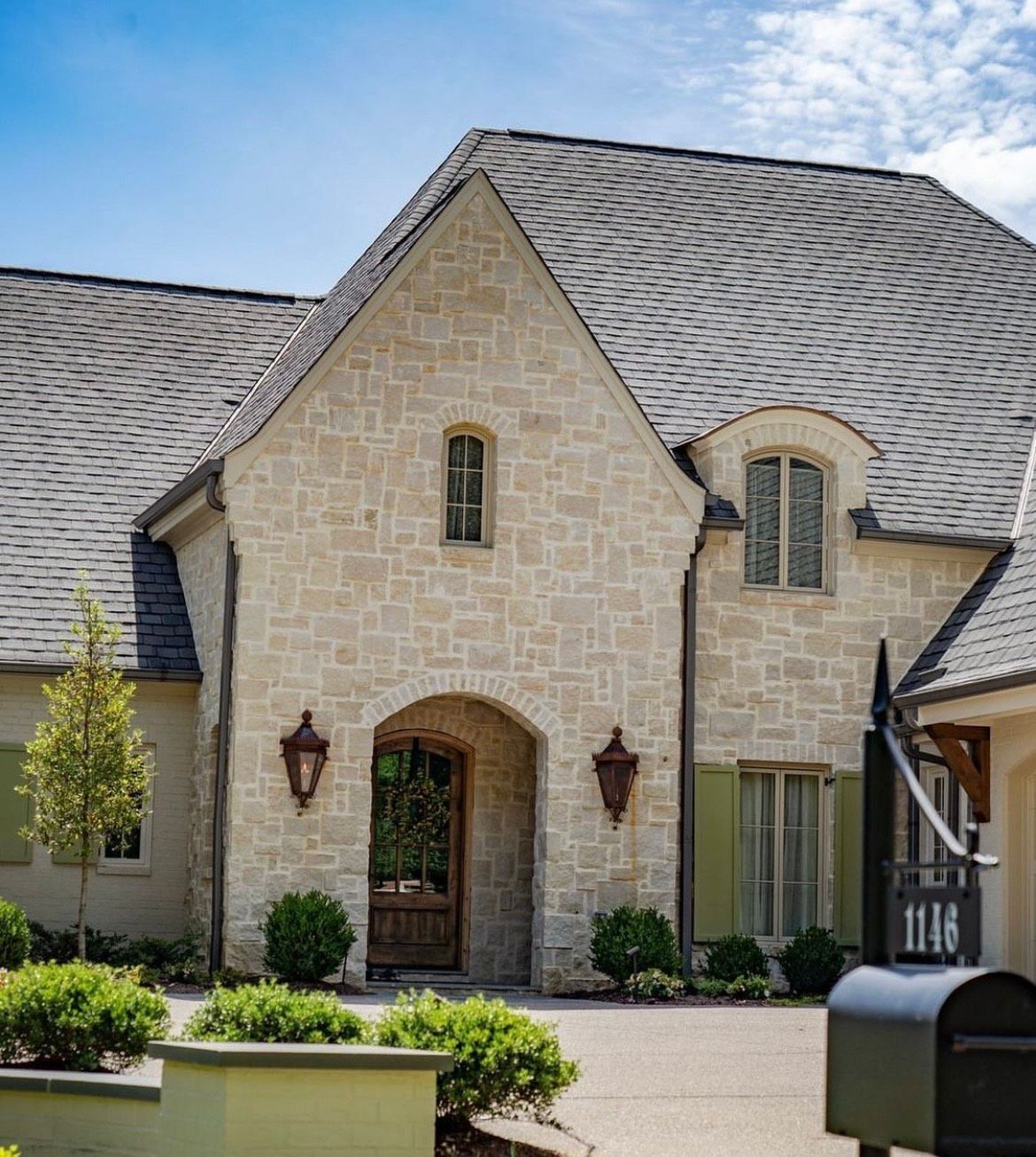 Stone house with arched doorway, two sconces, green shutters, and mailbox; a sunny day.