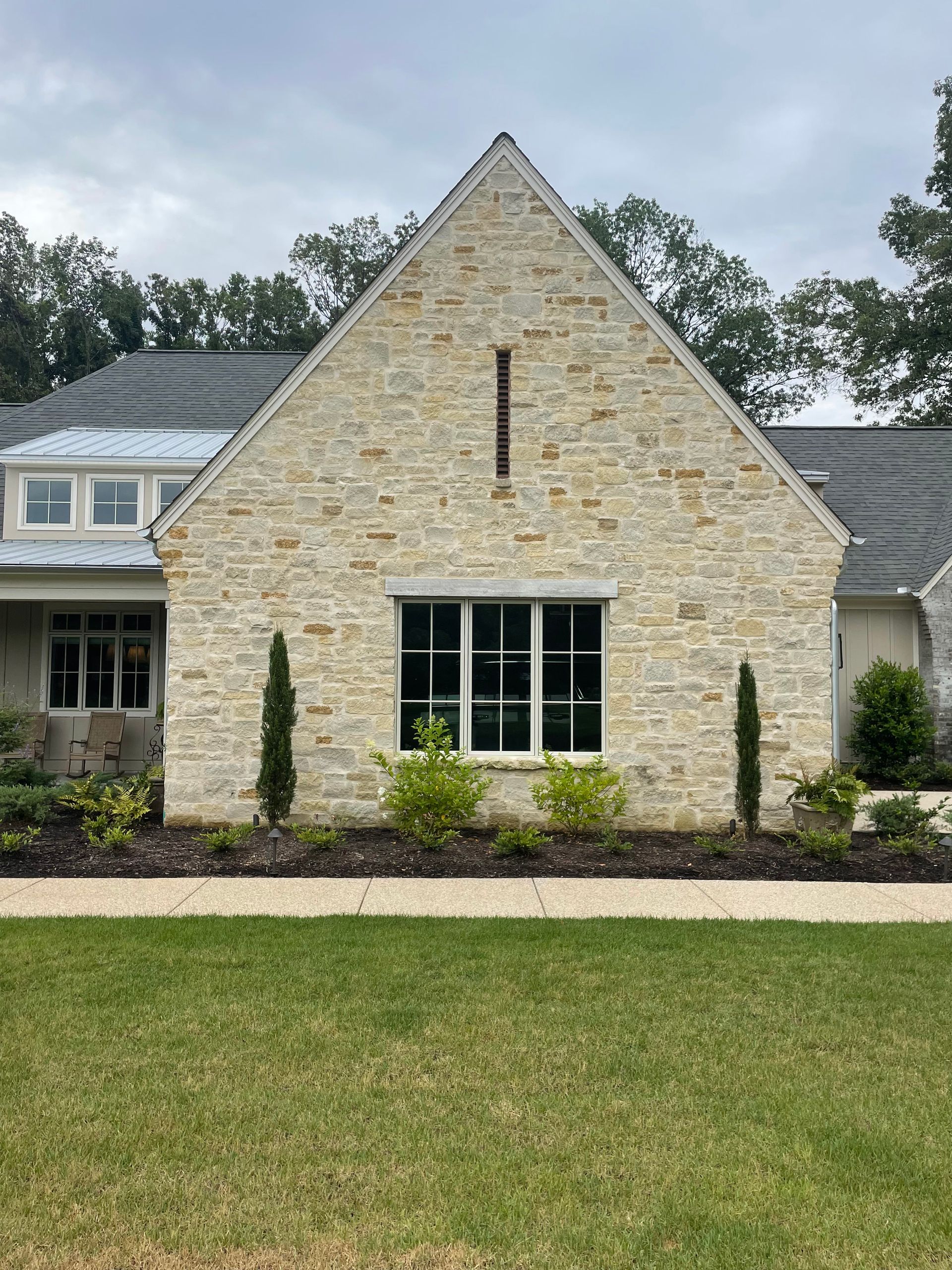Stone house with a gabled roof and a window on the facade. Two small trees flank the window and grass below.